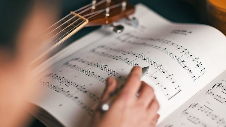繁體中文 (Traditional Chinese): 音樂系學生手持筆在五線譜本上進行作曲與和聲學理論分析，適合計劃海外留學或透過非聯招（Non-JUPAS）報讀英國、美國及香港頂尖音樂學院與大學音樂學位（Music）的同學參考。 簡體中文 (Simplified Chinese): 音乐系学生手持笔在五线谱本上进行作曲与和声学理论分析，适合计划海外留学或透过非联招（Non-JUPAS）报读英国、美国及香港顶尖音乐学院与大学音乐学位（Music）的同学参考。 英文 (English): A music student using a pen to compose and analyze music theory on a sheet music book, useful for students preparing portfolio auditions for top UK, US, and Hong Kong music schools.