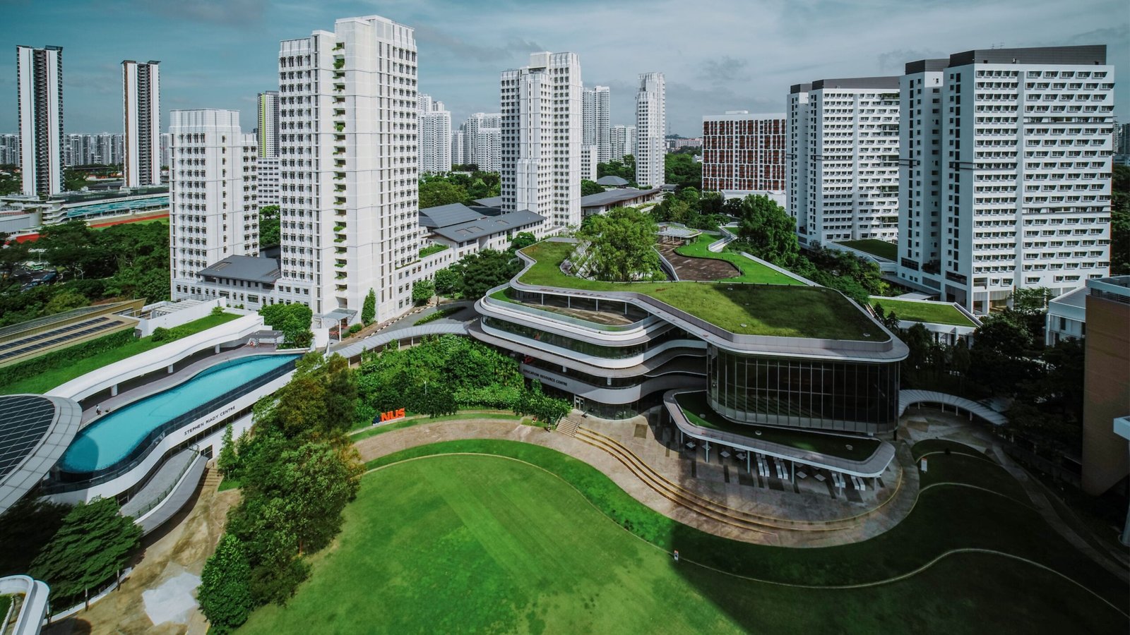 繁體中文 (Traditional Chinese)
鳥瞰新加坡國立大學（NUS）大學城（UTown）校園，畫面呈現標誌性的大草坪、極具現代感且覆蓋綠植屋頂的教學綜合大樓、露天游泳池以及高聳的學生宿舍群，展現世界級亞洲頂尖學府的國際化校園環境。簡體中文 (Simplified Chinese)
鸟瞰新加坡国立大学（NUS）大学城（UTown）校园，画面呈现标志性的大草坪、极具现代感且覆盖绿植屋顶的教学综合大楼、露天游泳池以及高耸的学生宿舍群，展现世界级亚洲顶尖学府的国际化校园环境。英文 (English)
An aerial drone view of the National University of Singapore (NUS) University Town (UTown) campus, featuring the iconic green lawn, modern eco-friendly buildings with rooftop gardens, an outdoor swimming pool, and student residential colleges under a bright sky.